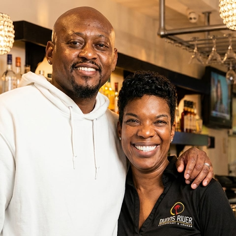 Smiling man and woman, likely owners, standing in front of a bar with bottles and glasses.