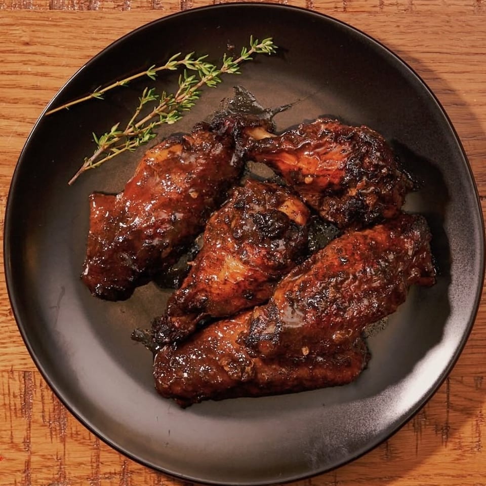 Four glazed chicken wings with a sprig of thyme on a black plate.