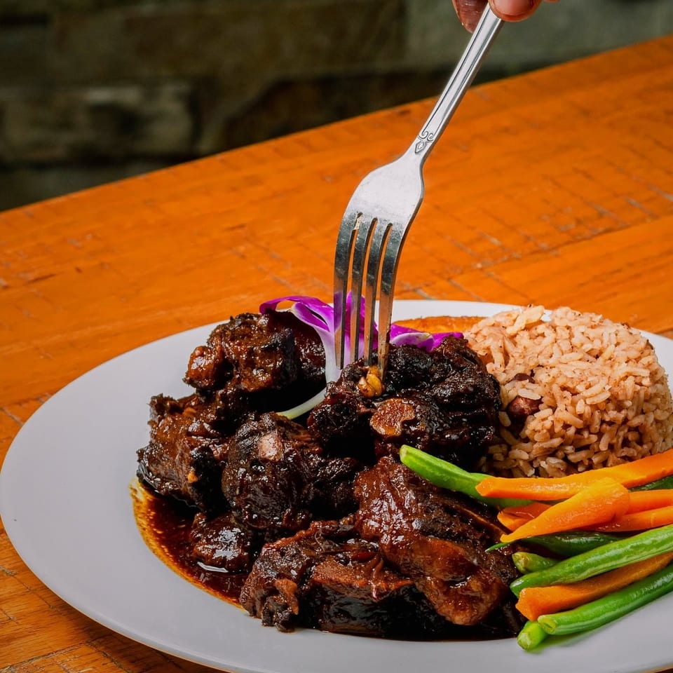 A hand uses a fork to pick up a piece of oxtail from a plate with rice, carrots, and green beans.