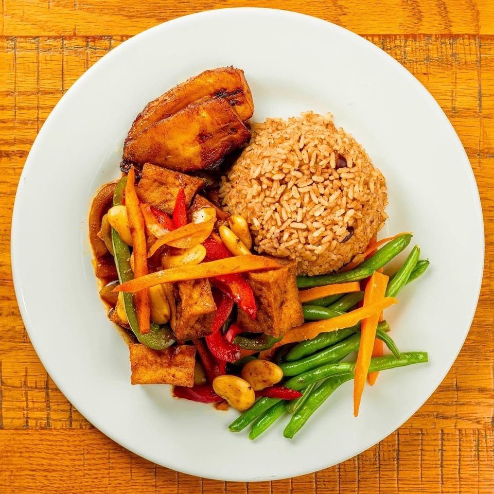 A white plate with fried plantains, rice, tofu stir-fry, green beans, and carrots.